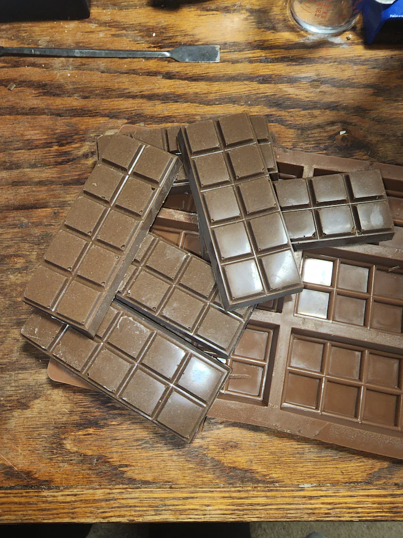 Several molded chocolate bars stacked on a wooden table next to a silicone mold and a metal tool, brown and tan tones